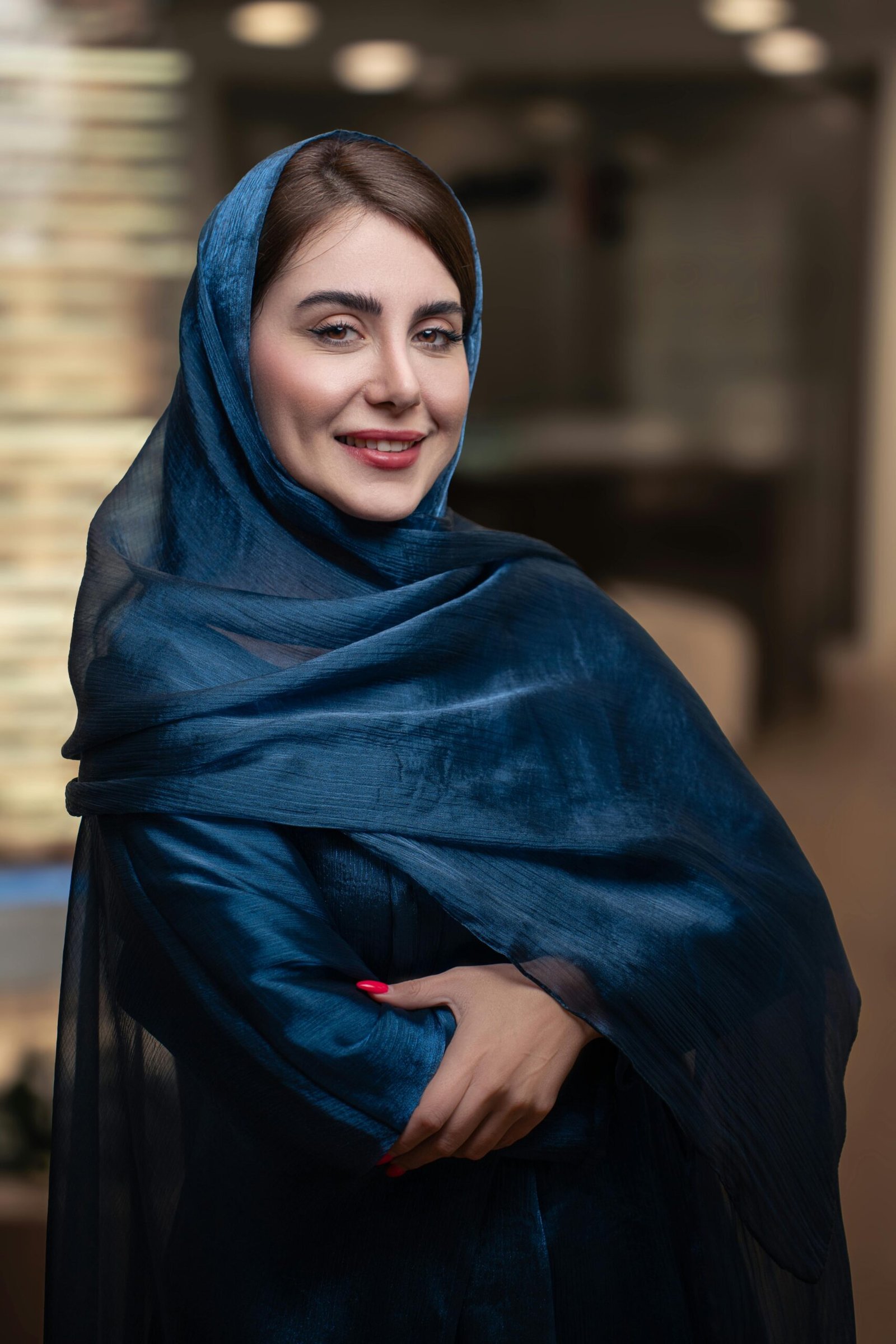A beautiful woman in a blue hijab smiles confidently indoors, showcasing elegance and cultural fashion.